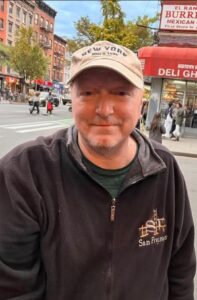 Dennis Tassone, man in a dark-colored hoodie stands in front of a street. He's wearing a tan cap.