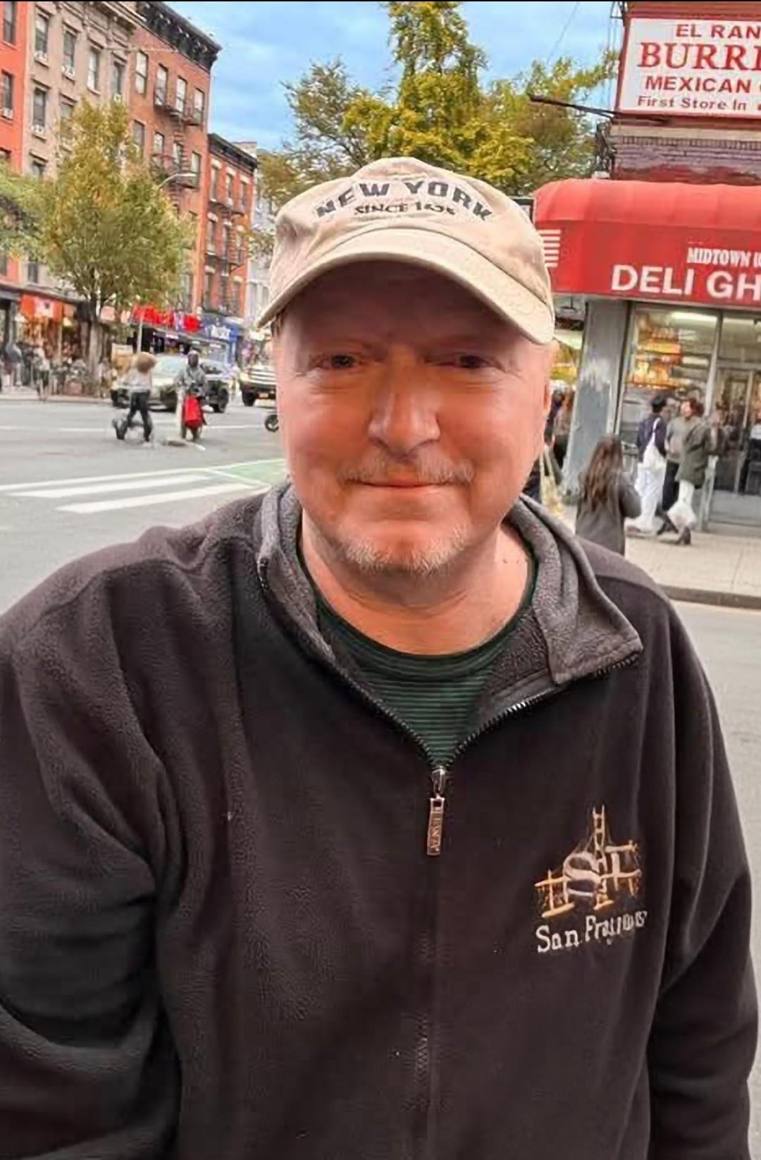 Dennis Tassone, man in a dark-colored hoodie stands in front of a street. He's wearing a tan cap.